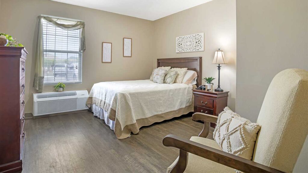 A cozy assisted living bedroom with a bed, white bedding, wooden headboard, nightstand with lamp, armchair with cushion, dresser, wall art, and a window letting in natural light. The wooden floor adds warmth and comfort.