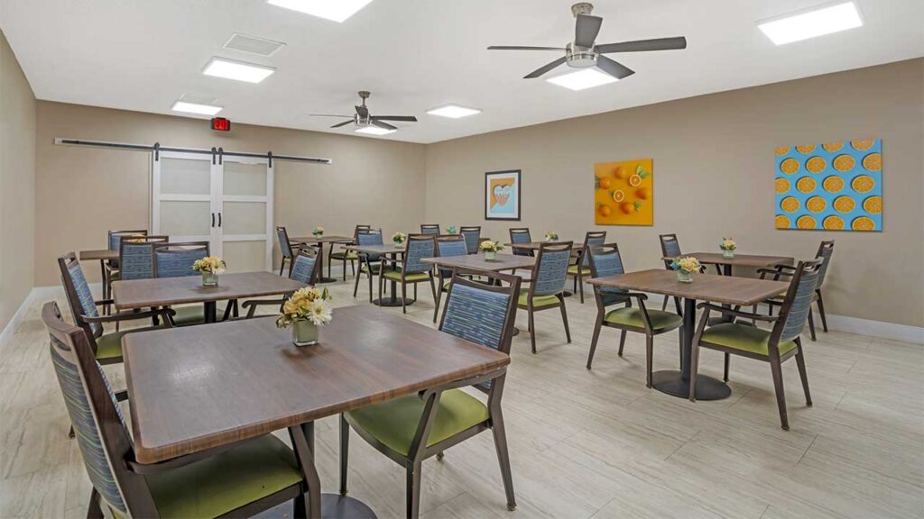 A modern dining room in an assisted living community features several square tables, each set with four chairs and small flower arrangements. Beige walls with colorful abstract artwork, ceiling fans, and bright lighting enhance the welcoming space.