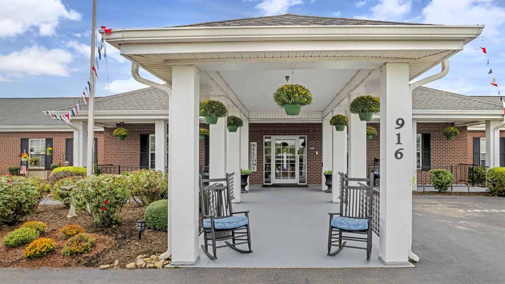 A covered entrance to a brick Assisted Living building with white columns, rocking chairs with blue cushions, and hanging plants. The address number 916 is visible on a column, while shrubs and flowers line the walkway.