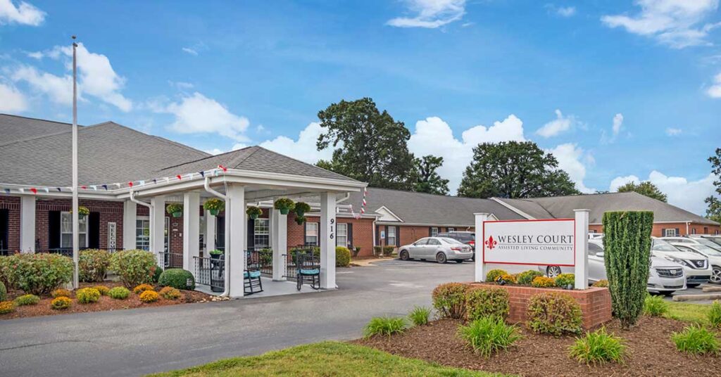 A single-story brick building with a covered entrance and rocking chairs sits beside a parking lot. A sign in front reads “Wesley Court Assisted Living Community.” The landscaped grounds, filled with flowers and shrubs, create a welcoming assisted living environment.