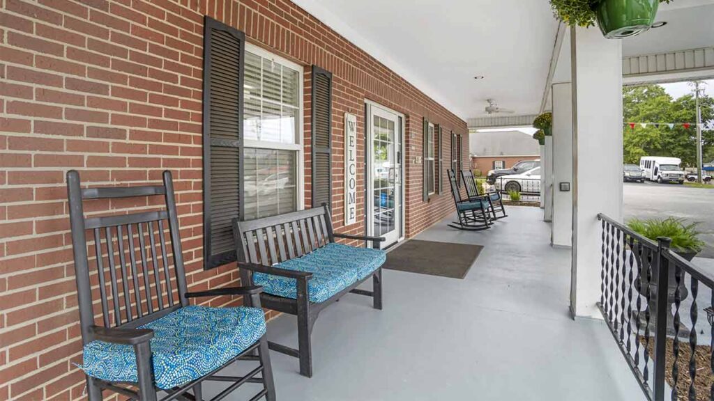 A wide, covered porch with a brick exterior features rocking chairs and benches with blue patterned cushions, black railings, and potted plants hanging near the ceiling—creating an inviting assisted living space.