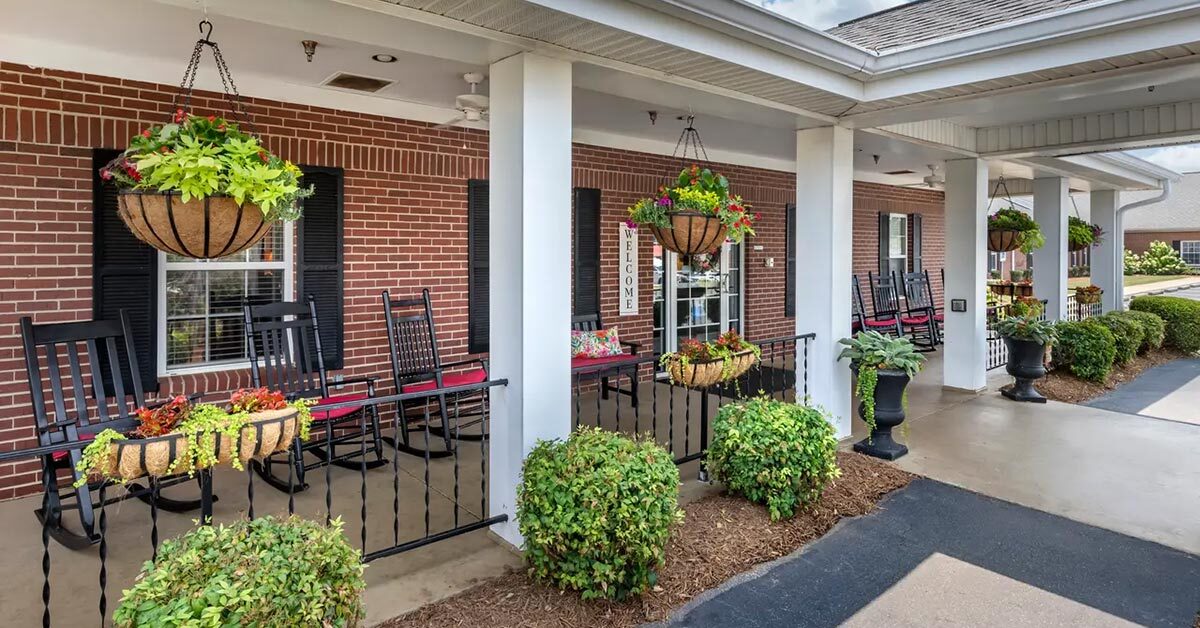 A covered porch with a brick exterior at the assisted living facility features multiple black rocking chairs adorned with red cushions. Hanging baskets filled with colorful flowers and plants decorate the area, while a paved walkway and manicured bushes line the foreground.