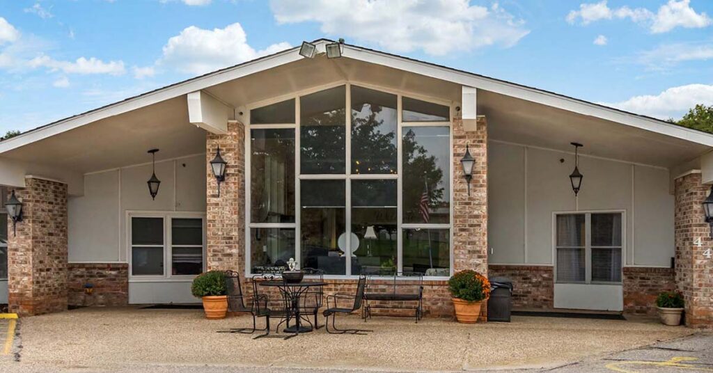 Single-story Rehabilitation & Health Care building with large floor-to-ceiling windows, brick and white siding exterior, sloped roof, outdoor seating with a black metal table and chairs, potted plants, and lantern-style lights by the entrance.