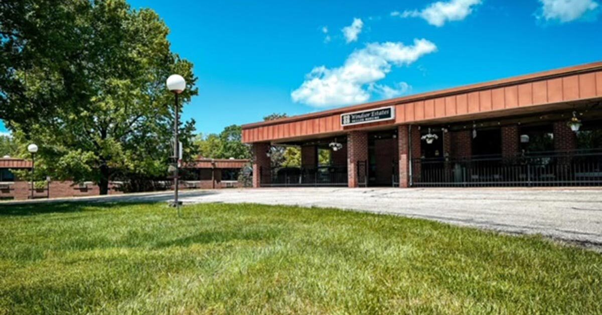 Exterior view of a long, single-story brick building with a brown roof, situated on a grassy area. The entrance to the senior rehabilitation center features a covered walkway with white light posts lining the path. Tall trees and a bright blue sky with a few clouds are in the background.