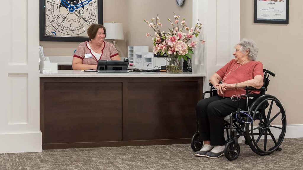 An elderly woman in a wheelchair is seated next to a reception desk, engaging in conversation with a receptionist who is smiling. The office is warmly decorated with a large flower arrangement and a wall clock.