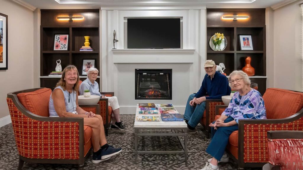 A group of four older adults sit in a cozy living room with orange chairs and a fireplace. Shelves with books and decor frame the fireplace, and magazines are spread out on the coffee table. They appear to be in conversation and enjoying each other's company.