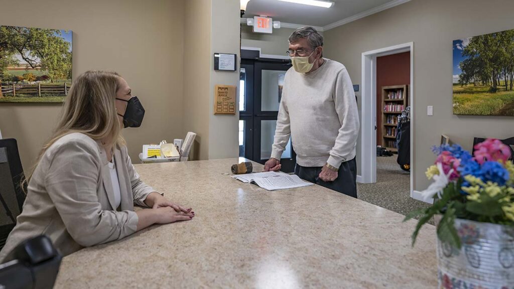 A man in a white sweater and face mask stands at a reception desk, speaking to a seated woman wearing a face mask and beige blazer. The office setting features paintings of landscapes and a shelf with books and items. A bouquet of flowers is in the foreground.