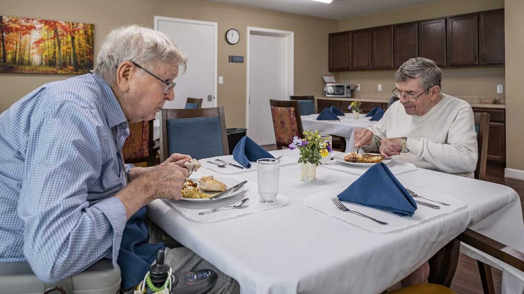 Two elderly men are seated at a dining table in a well-lit room with wooden cabinets and wall art. They are enjoying a meal together, using silverware and blue napkins are neatly placed on the table. A vase with flowers is in the center of the white tablecloth.