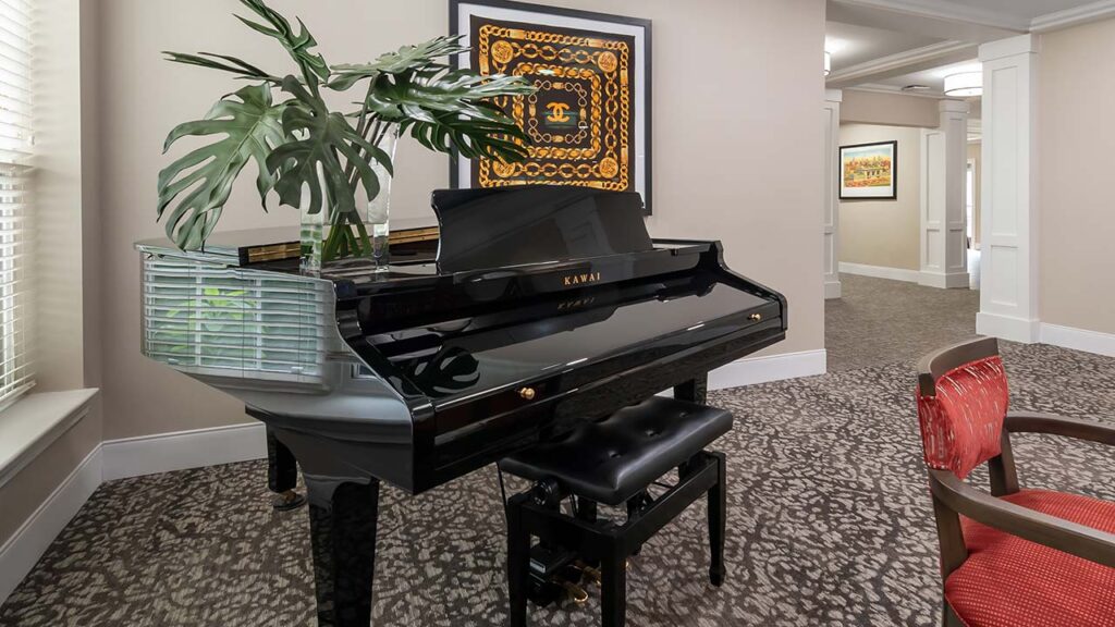 A black Kawai grand piano with a plant arrangement and a decorative framed artwork on the wall behind it is situated in a carpeted room. Nearby, there are light-colored walls, white trim, and a hallway leading to another area. A red chair is partially visible on the right.