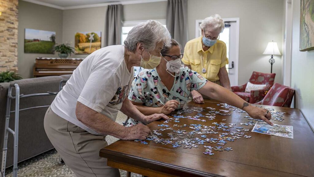 Three elderly individuals work on a jigsaw puzzle at a wooden table. Two women and a man, all wearing masks, focus on fitting pieces together. The room has a cozy atmosphere with a stone wall, piano, red armchairs, a lamp, and framed pictures.