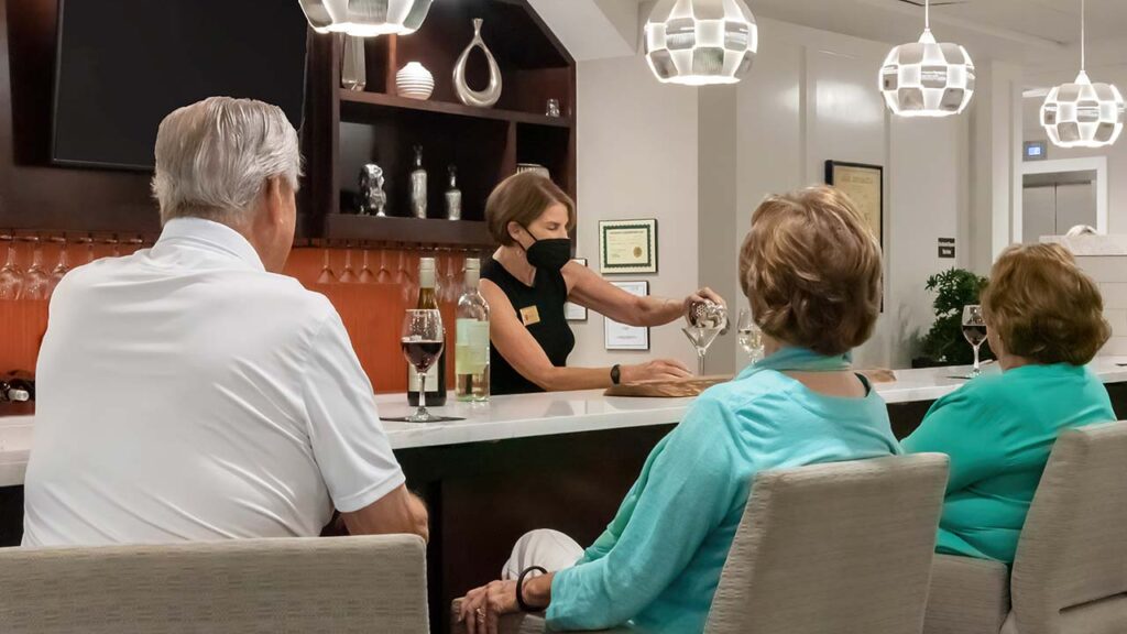 A bartender in a black mask serves white wine to three elderly patrons seated at a modern bar. The bartender is opening a bottle while the patrons watch. In the background, shelves with decorative items and glasses, along with pendant lights, are visible.