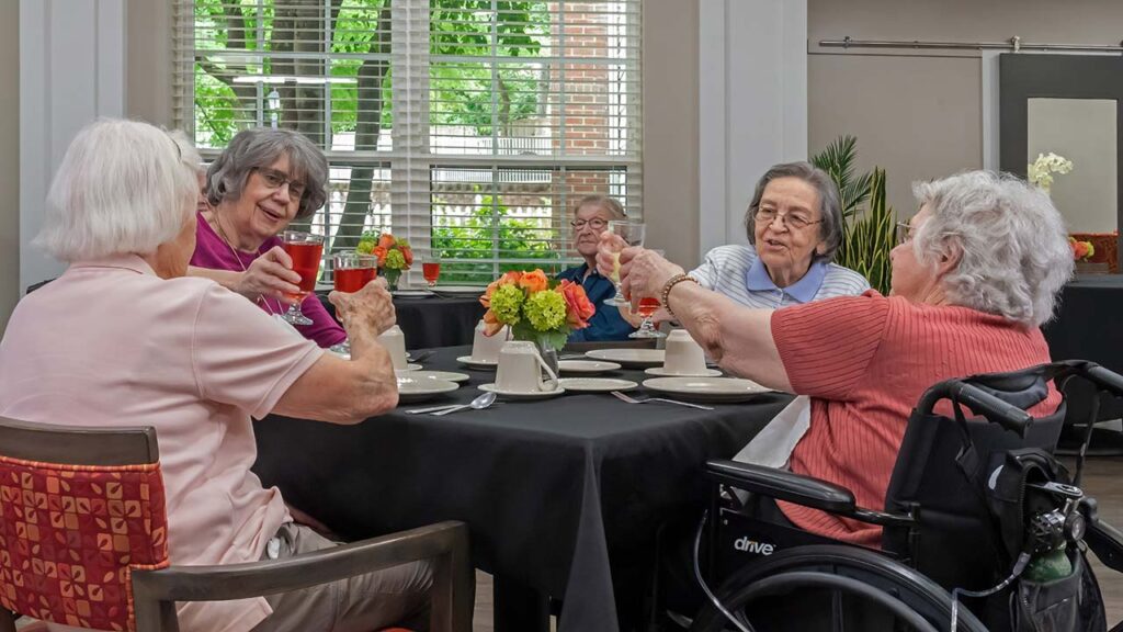 A group of five elderly women sit around a table in a brightly lit room with large windows, sharing a meal and raising their glasses in a toast. The table is set with plates, cups, and a vase of colorful flowers. One woman is in a wheelchair.