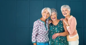 Three elderly women with gray hair stand closely together, smiling and laughing against a dark blue, paneled background. Dressed in casual, colorful tops, they share a joyful moment of friendship that perfectly captures the essence of senior living.