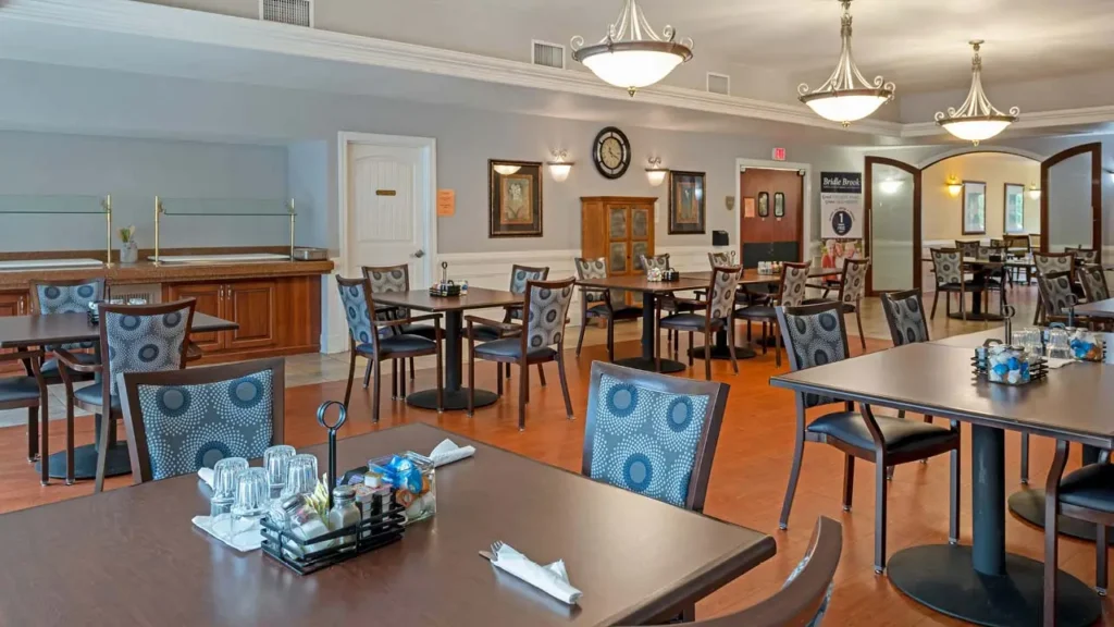 A spacious dining room features multiple tables and chairs arranged neatly. The tables are set with cutlery, condiments, and small centerpieces. The room has elegant chandeliers, wooden flooring, and a buffet counter on one side. The walls are painted light blue.