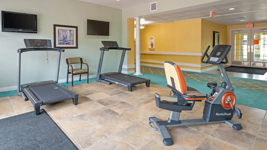A small, bright gym features two treadmills and an exercise bike on a tiled floor. The walls are painted yellow with framed pictures and a flat-screen TV mounted above, a brown chair, and a glass door leading to another room is visible on the right.