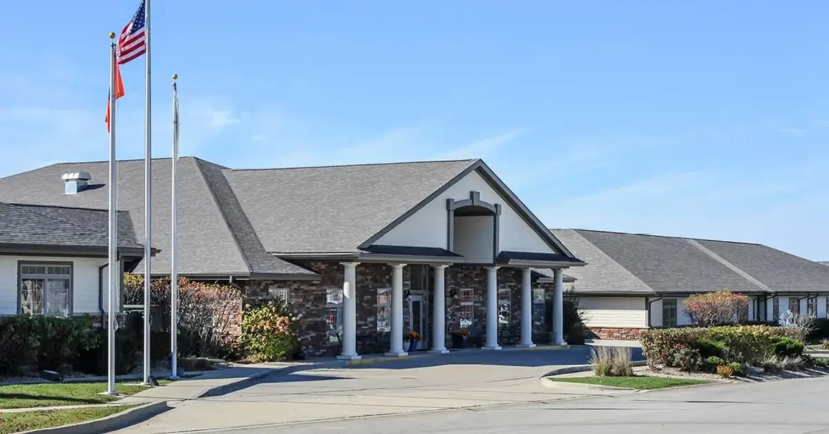 A single-story building with a stone facade, a gabled roof, and a covered entrance supported by columns. Two flagpoles with American flags stand in front. Surrounding shrubs and clear blue sky in the background.