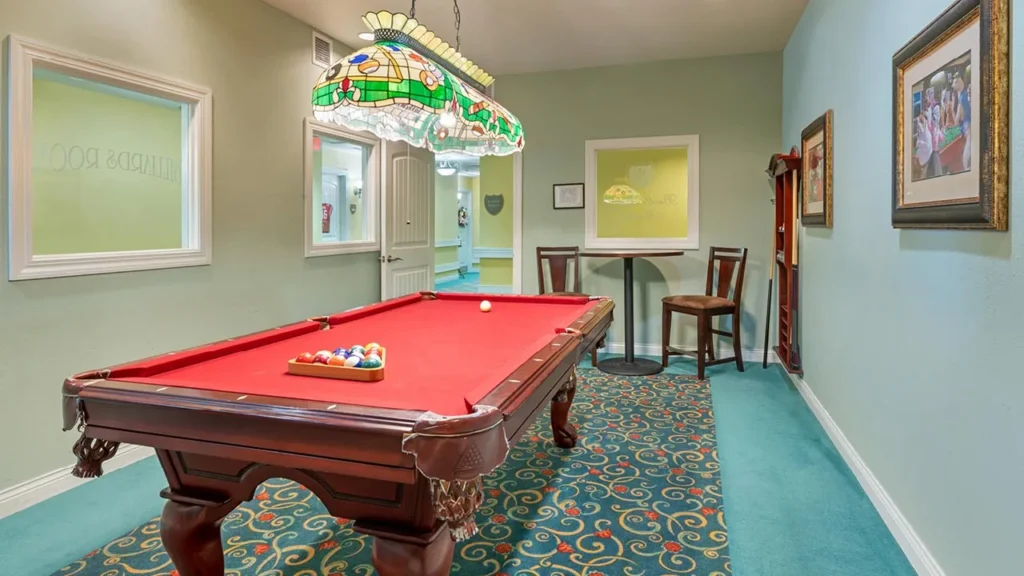 A room with a red-felt pool table under a colorful stained glass lamp. There is a rack with cues on the right wall, a high table with two chairs in the corner, and framed art on the walls. The carpet has a blue and gold pattern.