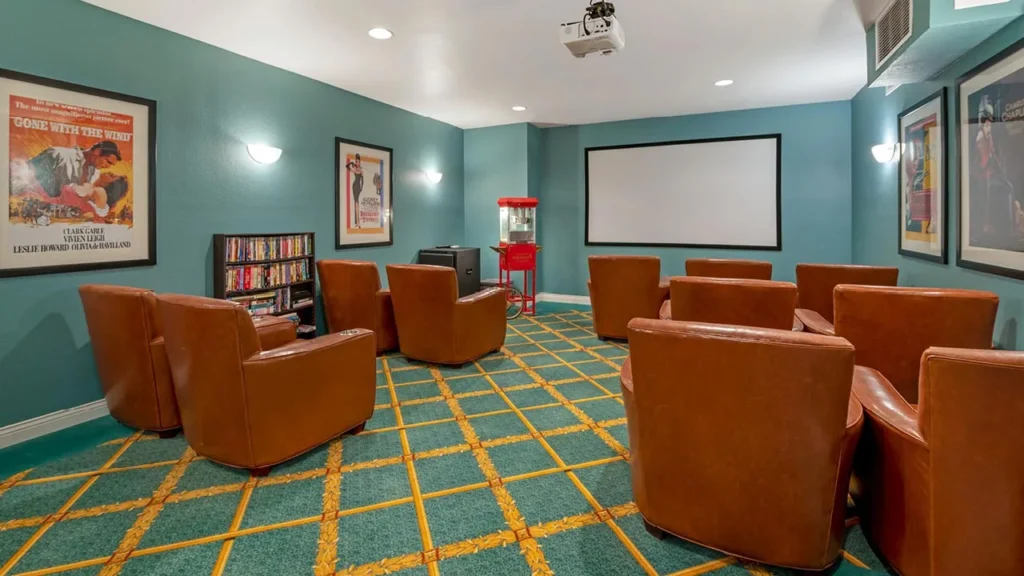 A small home theater room with eight brown leather armchairs facing a large white projector screen. The room has teal walls, movie posters, a popcorn machine, a DVD shelf, and yellow and green geometric-patterned carpet. Ceiling lights provide illumination.