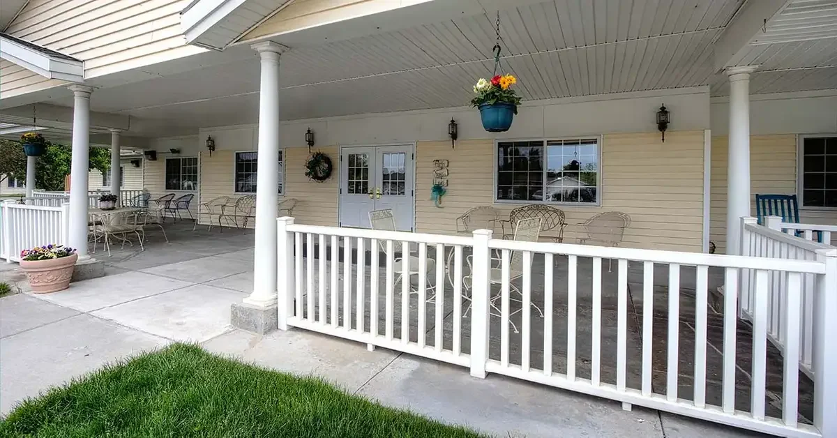 Outdoor patio area with white fencing and several round tables with chairs, each set beneath a covered porch. Hanging flower pots add color to the space, which is adjacent to a building with white siding and large windows. A grassy lawn stretches out in front.