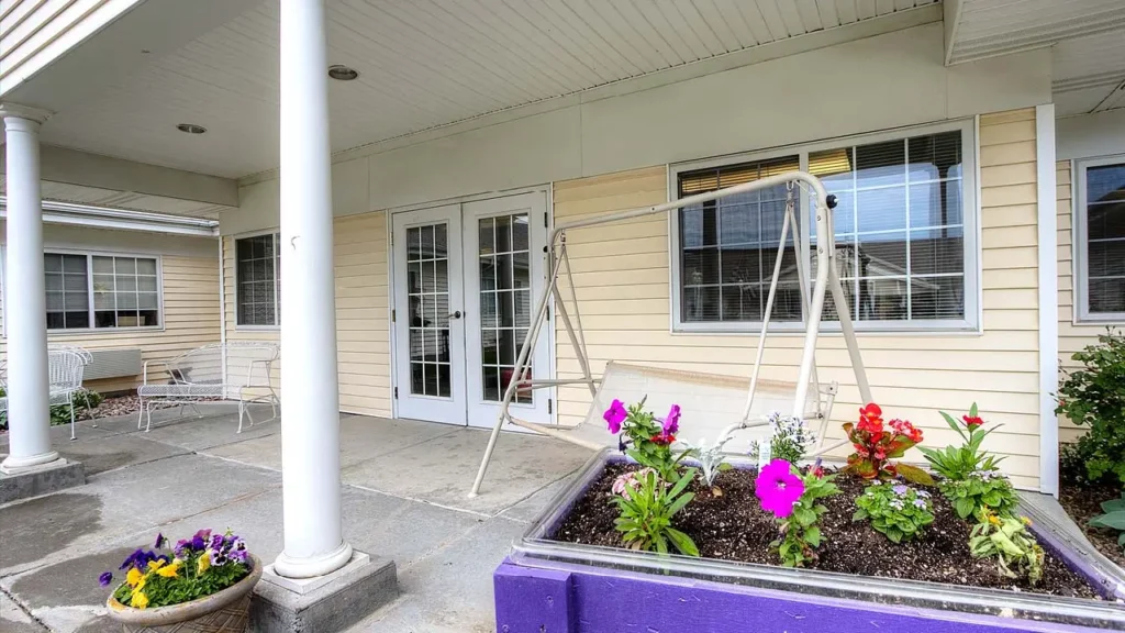 A quaint porch with a beige exterior, featuring a white swing set hanging from a metal frame. In the foreground, there's a purple planter box with blooming flowers. To the left, there are two white benches against the wall. Large windows and a glass door are visible.