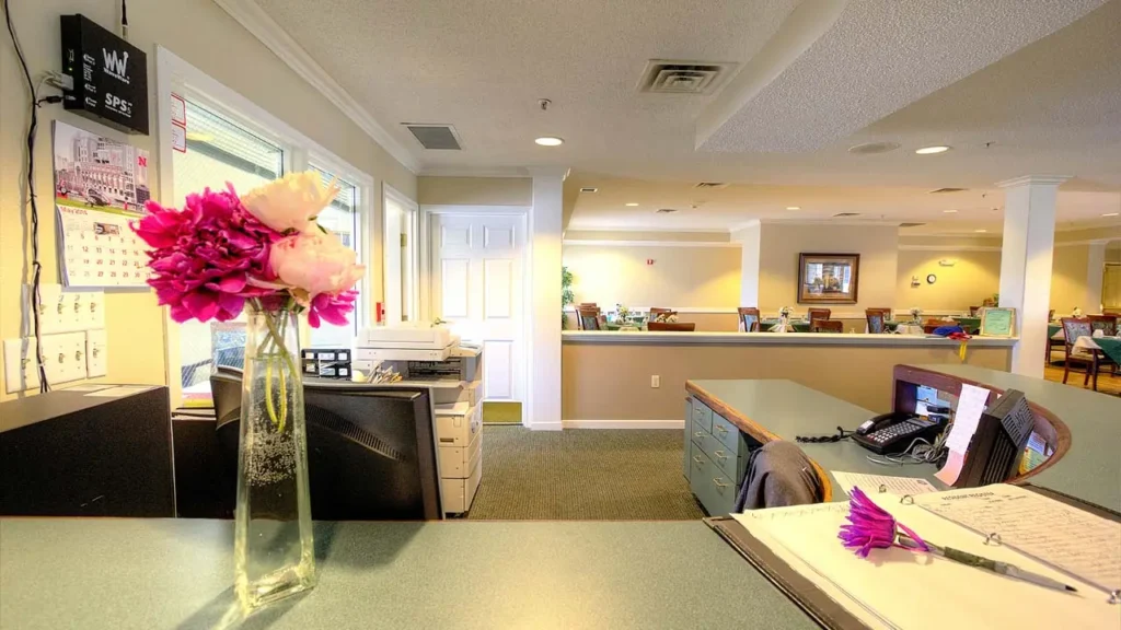 A brightly lit office reception area with a flower vase containing pink and white flowers on the counter. The counter also has a notebook, a pen, and an organizer. In the background, there are desks, chairs, and a few people working.