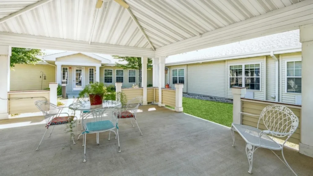 A covered patio with a white metal roof features a small round glass table with four white metal chairs. A red flower pot with greenery sits on the table. The patio is in a yard with green grass, and a light-colored building with multiple windows is visible in the background.