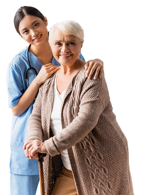 A young nurse in blue scrubs with a stethoscope around her neck is smiling and standing behind an elderly woman with short white hair, wearing a beige knitted cardigan, holding a cane. Both women are smiling and appear to be happy in their senior living environment.