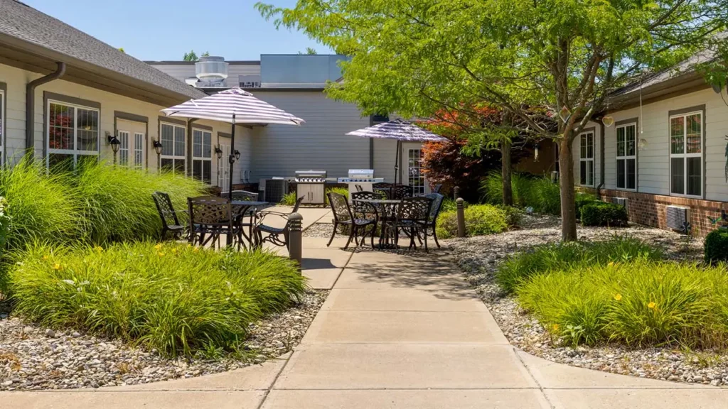 A courtyard with outdoor seating, featuring several black metal tables and chairs under striped purple umbrellas. The area is surrounded by lush green bushes and trees. The pathway is flanked by buildings with patio doors leading into them. The sky is clear and blue.