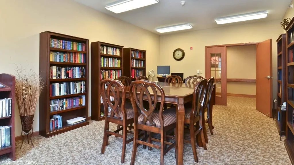 A cozy study room with wooden bookshelves filled with books lining the walls. A rectangular table with chairs occupies the center, and a doorway with a glass panel is visible on the right. A wall clock and a computer monitor are placed on the back wall.