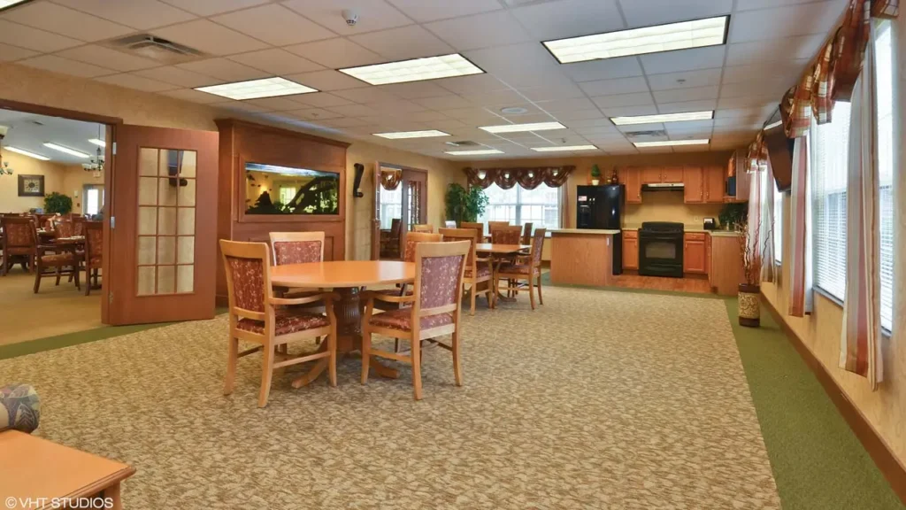 A spacious, well-lit communal dining area with round wooden tables and cushioned chairs. The room features large windows, a carpeted floor, and a kitchen in the background equipped with an oven, fridge, and sink. A large aquarium and wooden doors are visible on one side.