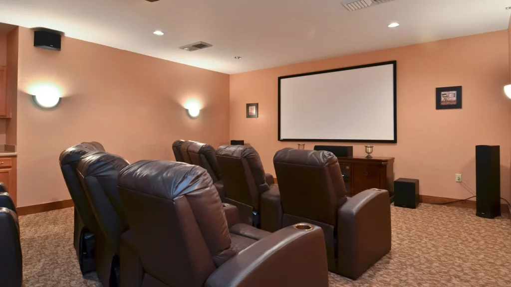 A home theater room with beige walls and carpeted floor. It features two rows of leather recliner chairs facing a large projection screen. Wall-mounted modern light fixtures provide ambient lighting. A console with speakers is placed below the screen.
