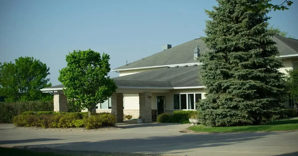 A suburban house with a sloped roof, stone and siding exterior, surrounded by lush greenery, including tall evergreens and manicured bushes. There is a paved driveway and a small front porch area flanked by shrubs. The sky is clear and blue.