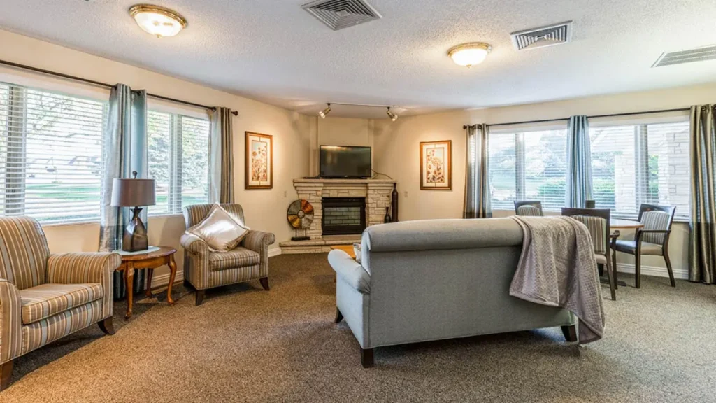 A cozy living room with carpeted floors, featuring a grey sofa with a blanket draped over it, two striped armchairs, a small round table with a lamp, and a dining area with a table and four chairs. A brick fireplace with a TV above it and two windows brighten the space.