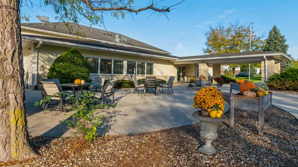 A spacious outdoor patio of a house featuring several tables and chairs. Seasonal decorations, including pumpkins and pots of yellow mums, adorn the area. A covered driveway is on the right, and the patio is surrounded by green bushes and a gravel path.