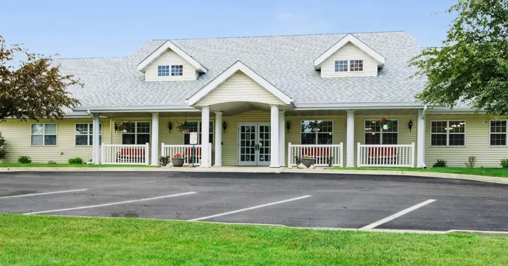 A single-story building with a grey gabled roof, cream-colored siding, and dormer windows. It has a covered entrance with white pillars, a white railing, and potted plants. The front area includes a paved driveway and a few parking spaces, with surrounding greenery.