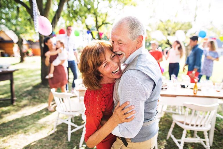 An older couple embraces and laughs while dancing outdoors at a festive gathering. People in the background socialize near a table with colorful decorations, creating a cheerful atmosphere under the trees.