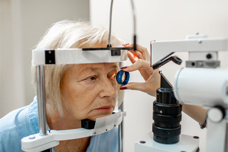 An elderly woman undergoes an eye examination. She is positioned at a slit lamp, an instrument used for eye testing. A healthcare professional, with red painted nails, is holding a blue lens close to her right eye. The woman looks forward attentively.