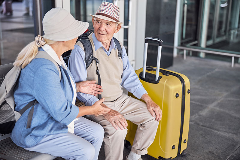 Senior couple with hats and yellow suitcase sitting and talking outside airport