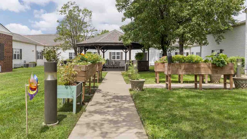 A serene garden area with raised wooden planters filled with greenery and flowers. A concrete pathway leads to a central gazebo surrounded by trees and grass. White buildings border the garden. Decorative elements, including a small windmill, enhance the pleasant ambiance.