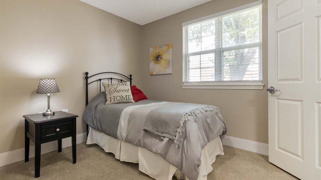 A bedroom with beige walls features a single bed with gray and white bedding and a red accent pillow. A black nightstand with a lamp is next to the bed. A window with white blinds, a floral painting, and an open door are also visible.