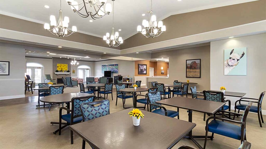 A spacious, well-lit dining area adorned with elegant chandeliers, square tables, and blue-patterned chairs. Walls feature colorful artwork and a counter is visible in the background. Small plants with yellow flowers decorate each table.