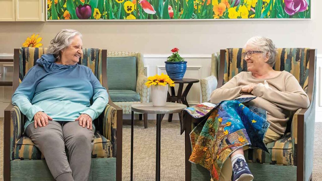 Two elderly women sit in armchairs facing each other, smiling and engaging in conversation. One woman holds a magazine. Behind them is a bright, colorful painting of flowers, and between them is a small table with a potted plant and a vase of sunflowers.