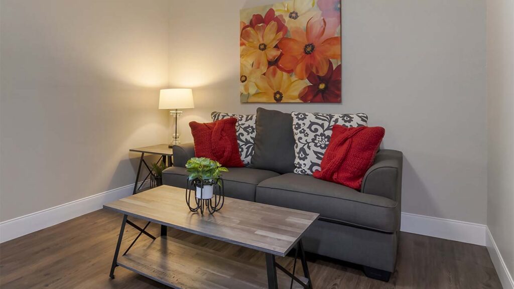 A cozy living room with a gray sofa adorned with red and patterned throw pillows. A wooden coffee table with a small potted plant is in front of the sofa. A side table with a lamp is beside the sofa, and a colorful floral painting hangs above it.