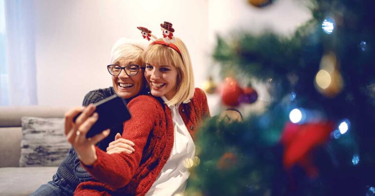 Senior woman and daughter wearing holiday accessories taking selfie by Christmas tree