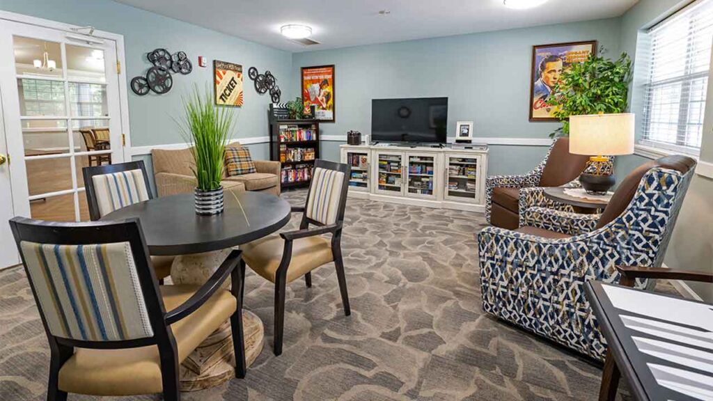 A cozy living room with a round table, four chairs with striped upholstery, two patterned armchairs, and a sofa. There's a TV on a white console, bookshelves, and movie posters on the walls. A large plant and a lamp add to the decor. Sunlight filters in through the windows.