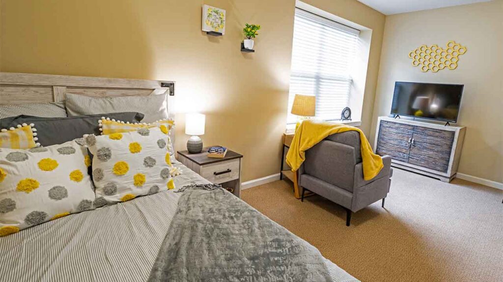 A cozy bedroom features a bed with a gray and yellow-themed bedding set, a small bedside table with a lamp, a comfortable gray chair with a yellow blanket draped over it, a flat-screen TV on a wooden dresser, and wall decor including a potted plant and hexagonal art.