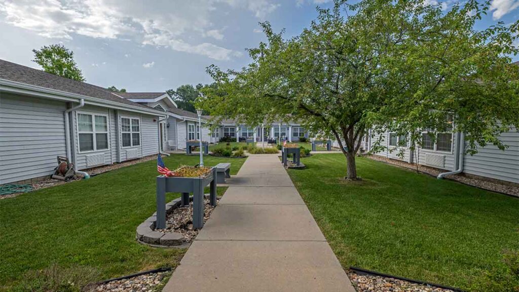 A well-maintained pathway flanked by green lawns leads to a series of single-story white buildings. Flower beds with American flags are positioned along the walkway, and a large tree provides shade in the center of the image under a partly cloudy sky.