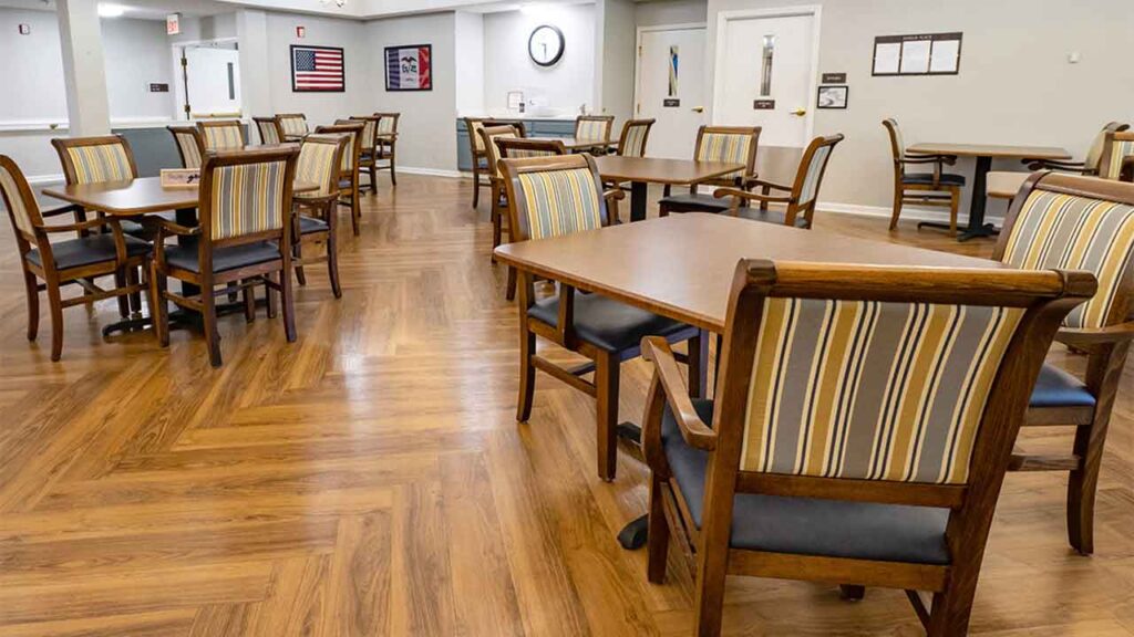 A spacious room features wooden dining tables and matching chairs with striped upholstery. The hardwood floor is arranged in a herringbone pattern. Walls are adorned with framed pictures, an American flag, and a clock. The space is brightly lit and well-organized.
