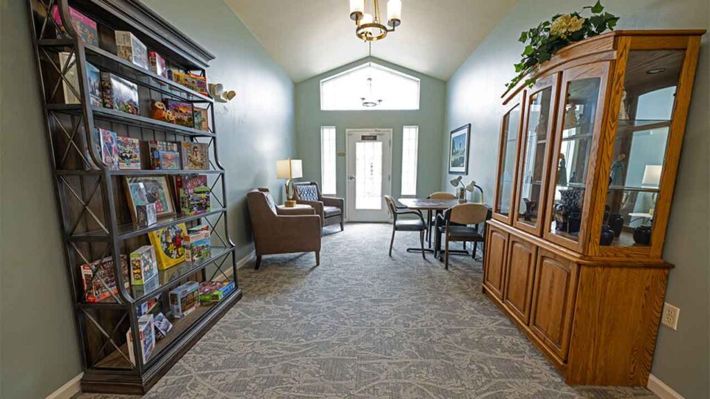A cozy room with a high ceiling and light blue walls features a bookshelf filled with colorful books on the left, a seating area with armchairs and a lamp at the back, and a wooden display cabinet with glass doors on the right. A door and windows are at the end.
