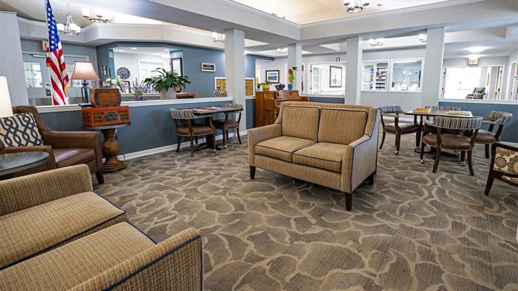 A spacious, well-lit room with beige carpeting and various seating arrangements, including armchairs and sofas. There's an American flag, potted plants, and a variety of tables and chairs. The walls are painted blue and white, and the room has a friendly, welcoming atmosphere.